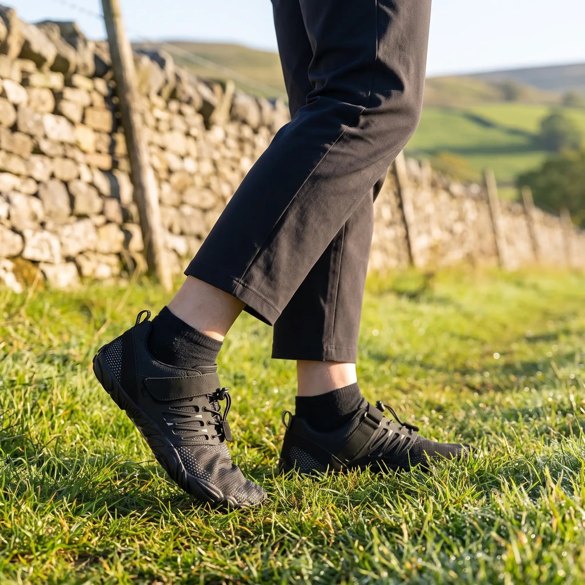 Black barefoot shoes worn on a grass footpath beside a dry-stone wall