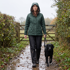 Woman walking a black Labrador on a wet muddy footpath in Britain in rain trousers and walking boots