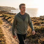 Man wearing the army green sun hoodie walking a coastal path at golden hour