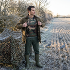Man shrugging a waxed jacket on over the camo base layer top beside a netting hide on a frosty stubble field in Britain