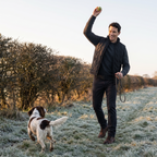 Man in a black body warmer throwing a ball for his spaniel in a frosty field in Britain
