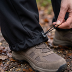 Close-up of a hand drawing the hem cord on the women's walking trousers, gathering the wide leg into a closed cuff over a walking boot