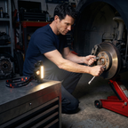 Head torch standing on its magnet end cap on a steel tool chest, lighting a brake disc while a man works with both hands