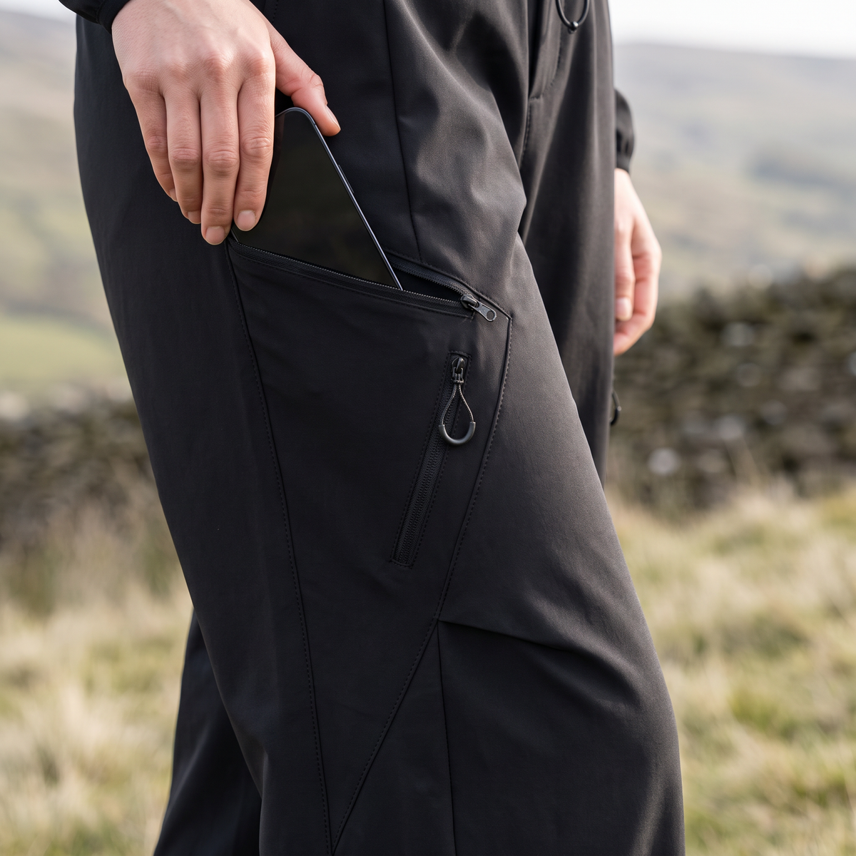 Close-up of a phone being put into the diagonal zip thigh pocket of the women's walking trousers, British hillside behind