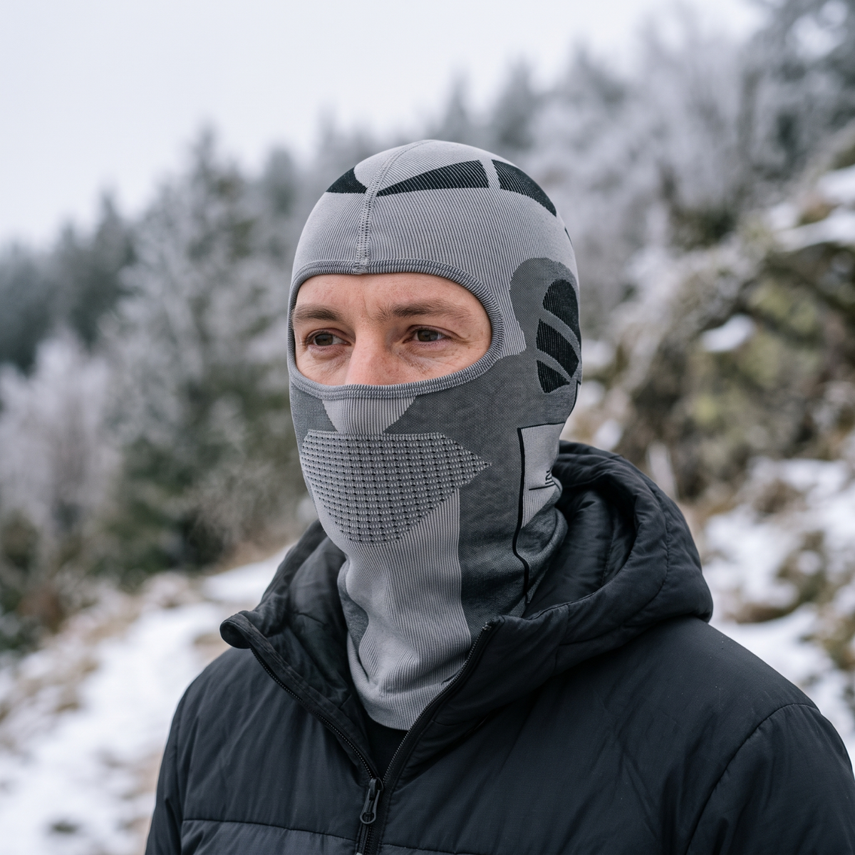 Man wearing the grey thermal balaclava under a jacket hood on a snowy hillside, face and neck covered