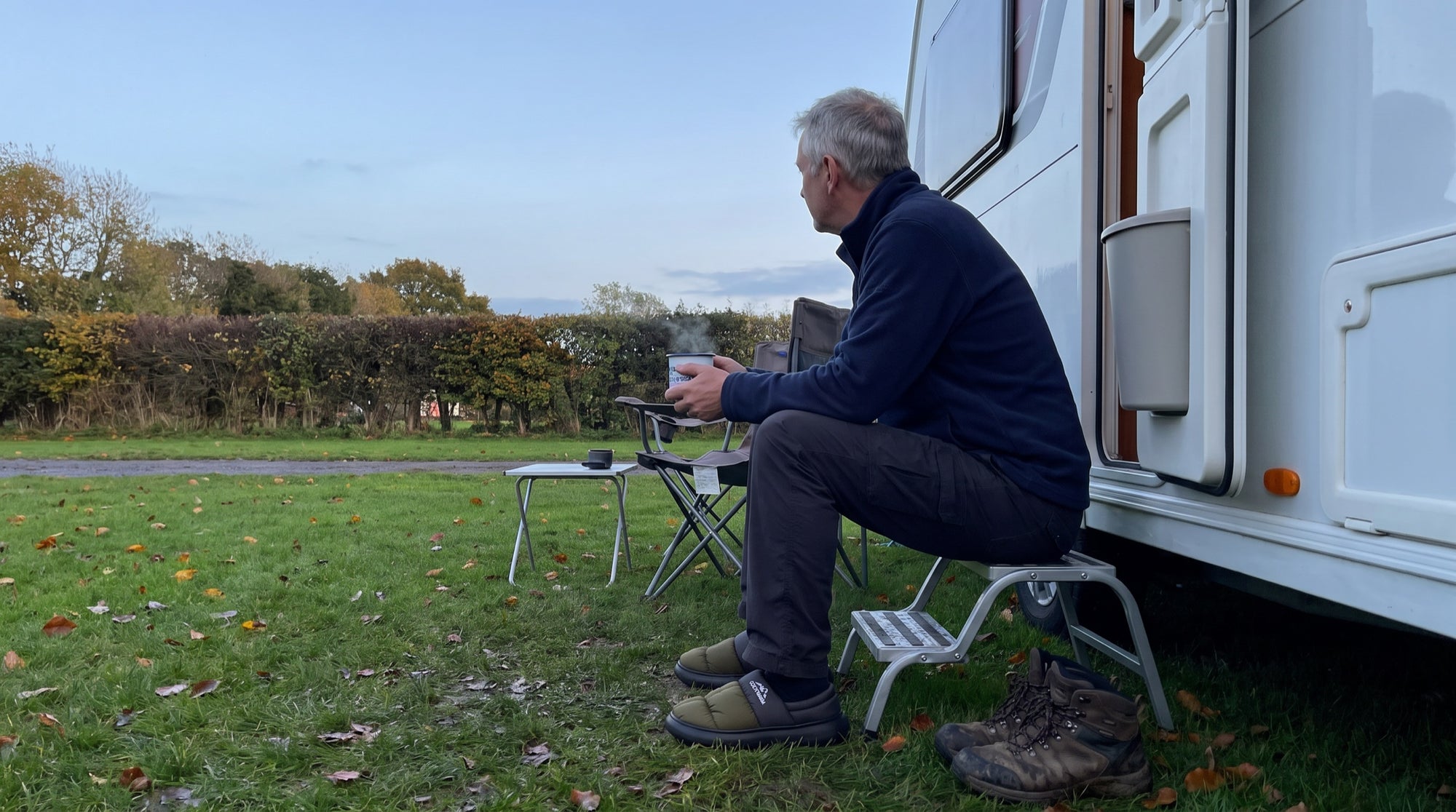 Camper sitting on a caravan step at dusk with a mug, wearing Terrabound Camp Slippers, walking boots left on the grass beside him