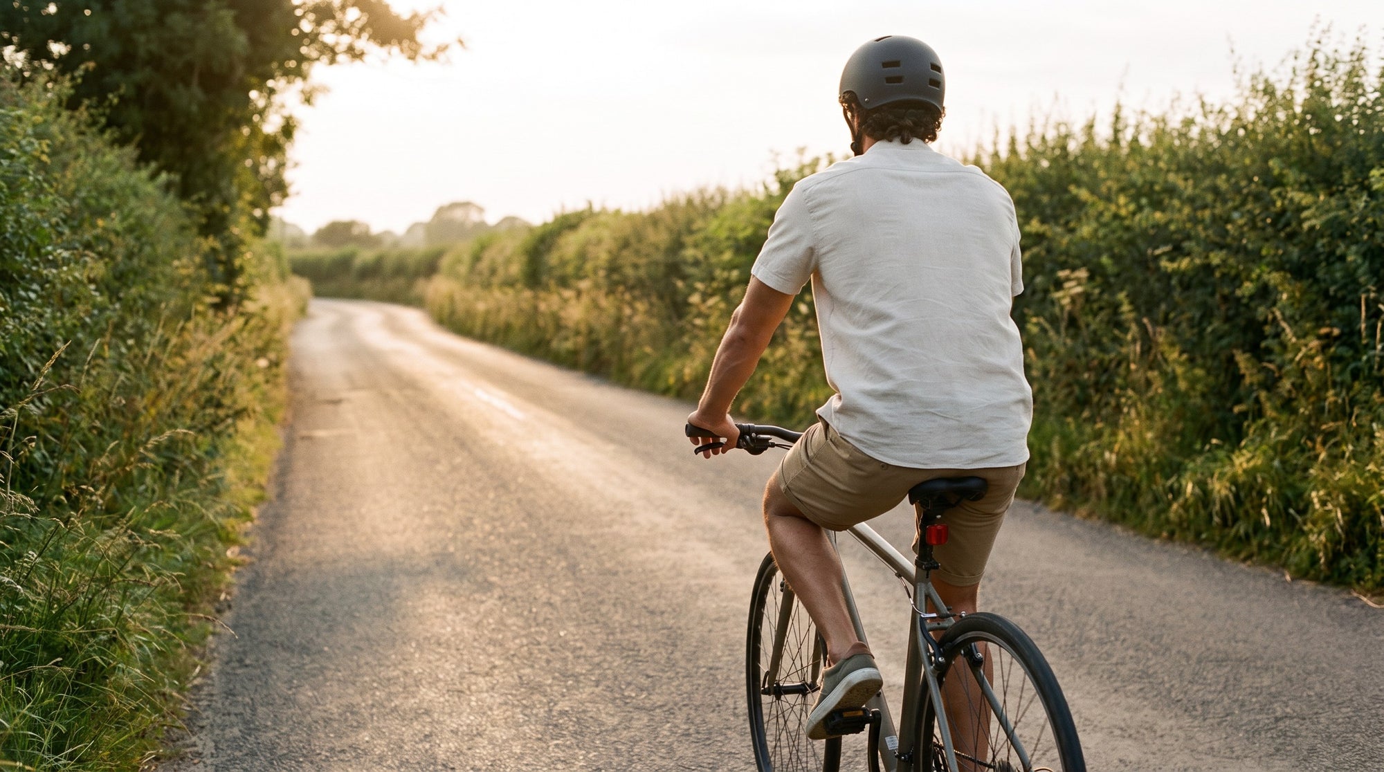 Man riding a bike down a quiet lane in his normal clothes, seen from behind in low evening sun