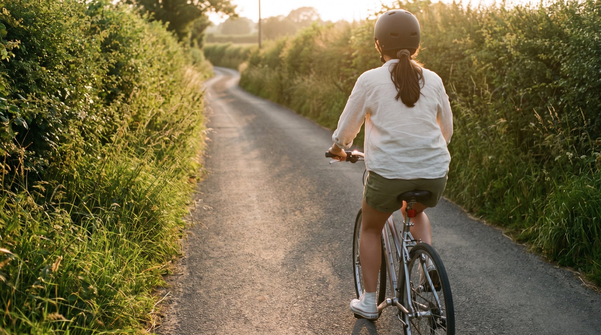Woman riding a bike down a quiet lane in her normal clothes, seen from behind in low evening sun