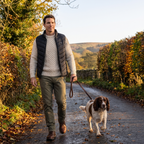 Man walking a spaniel on a country lane in autumn wearing the Terrabound men's padded gilet over a jumper