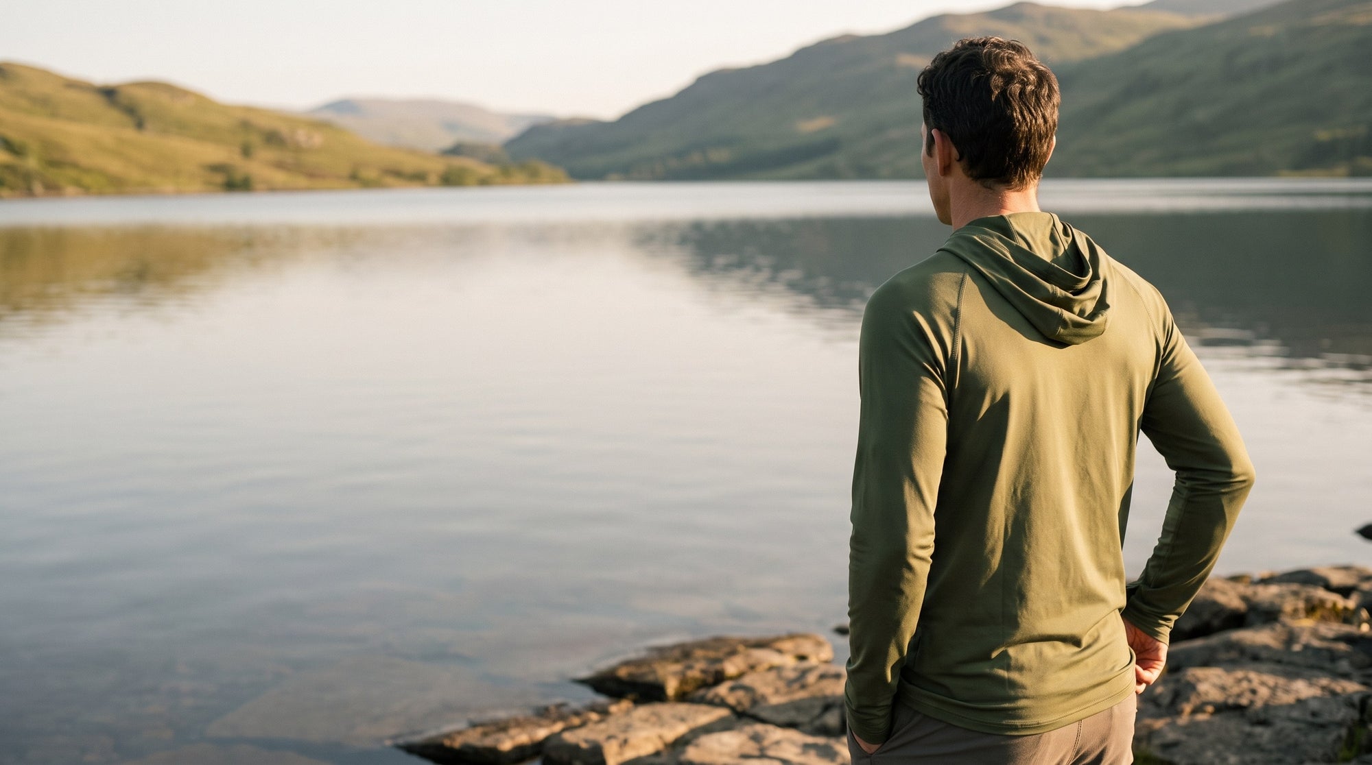 Man in the olive sun hoodie seen from behind, looking out across a loch in low evening sun
