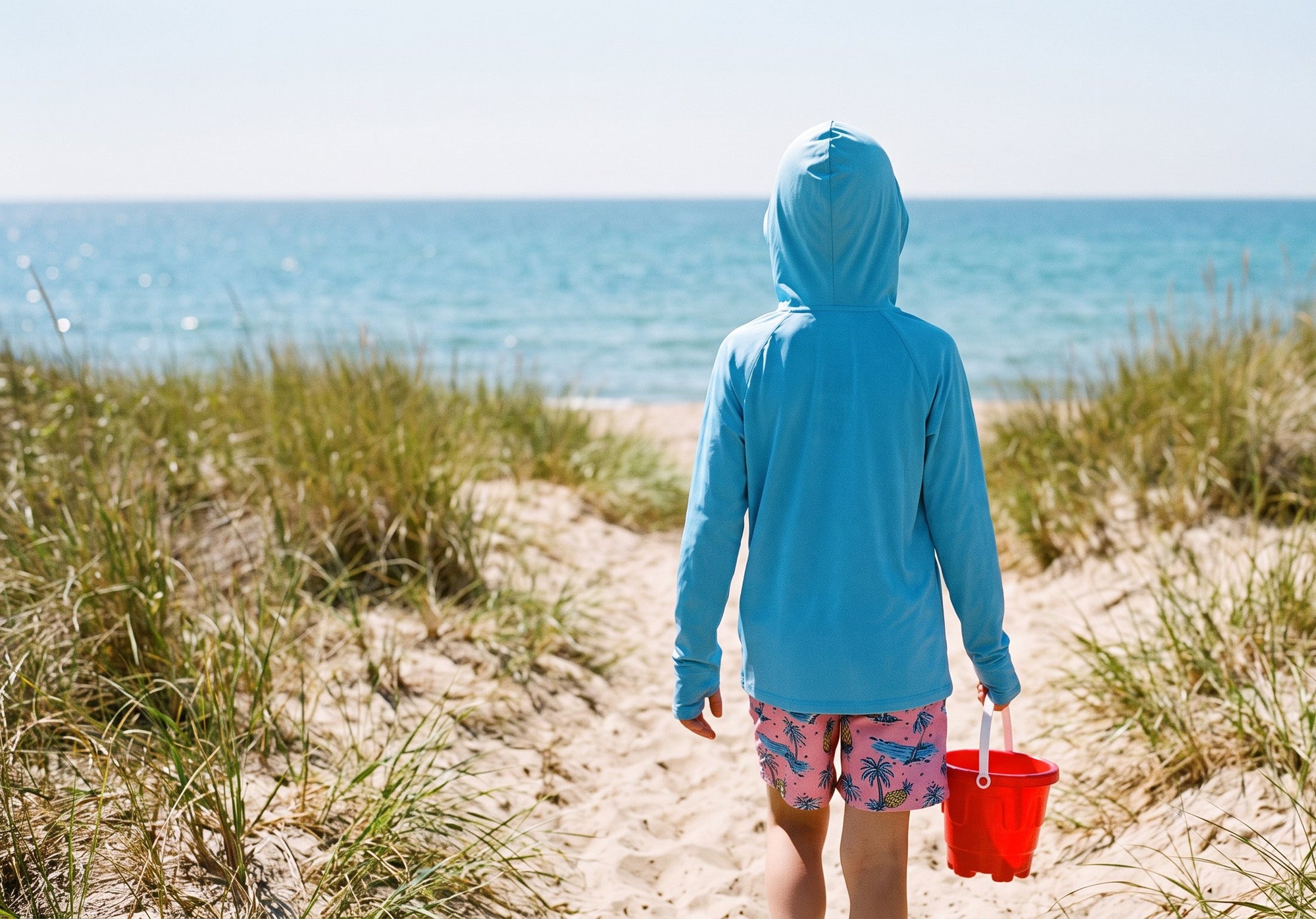 Boy in the sky blue Kids' Sun Hoodie seen from behind, hood up and thumbs through the cuffs, walking down a sandy path to the sea