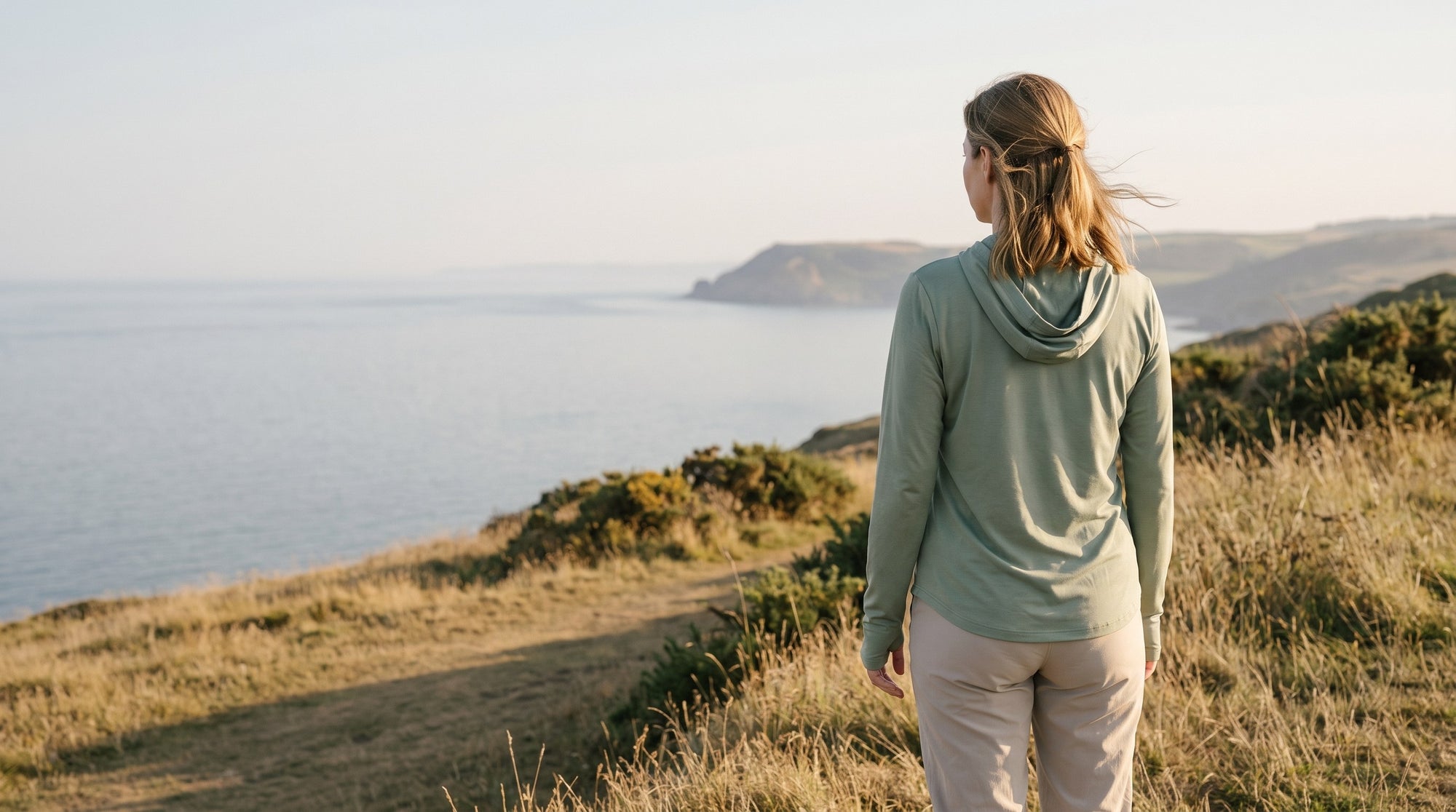 Woman in the sage sun hoodie seen from behind on a coast path, looking out over the sea in low evening sun