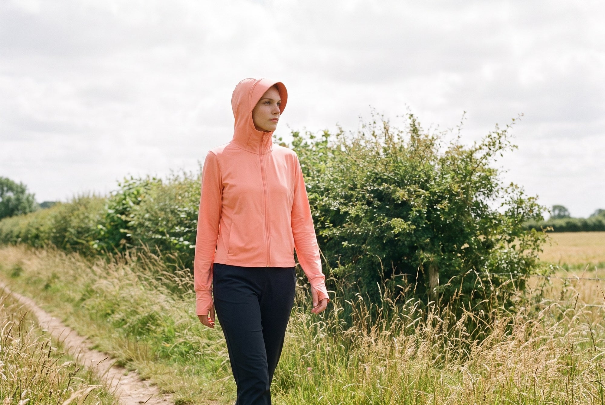 Woman walking a hedged field path in the coral Zip-Up Sun Hoodie with the hood up