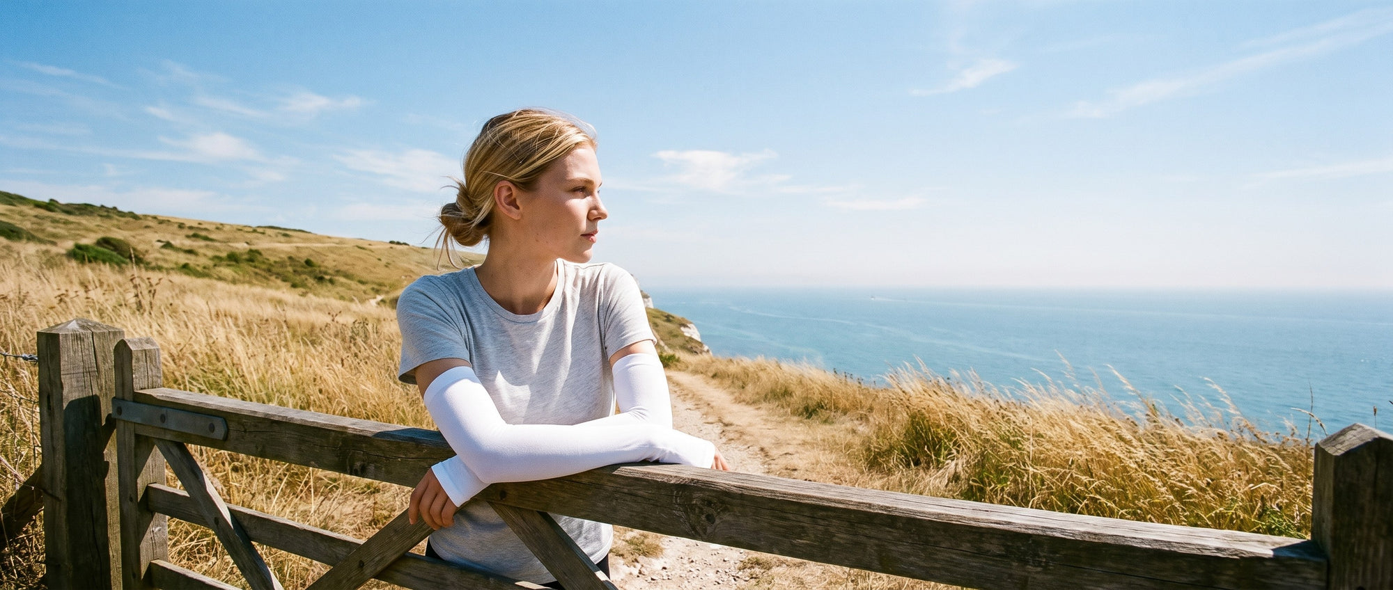 Woman resting her arms on a field gate above the sea in bright midday sun, wearing a short-sleeve t-shirt with white sun sleeves covering both arms