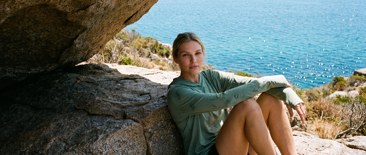 A woman in a long-sleeve sun shirt sitting on a granite ledge above a blue bay in bright sun